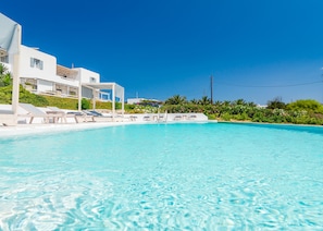 A clear blue pool with white lounge chairs and a white building in the background. - En Milo (Milos)