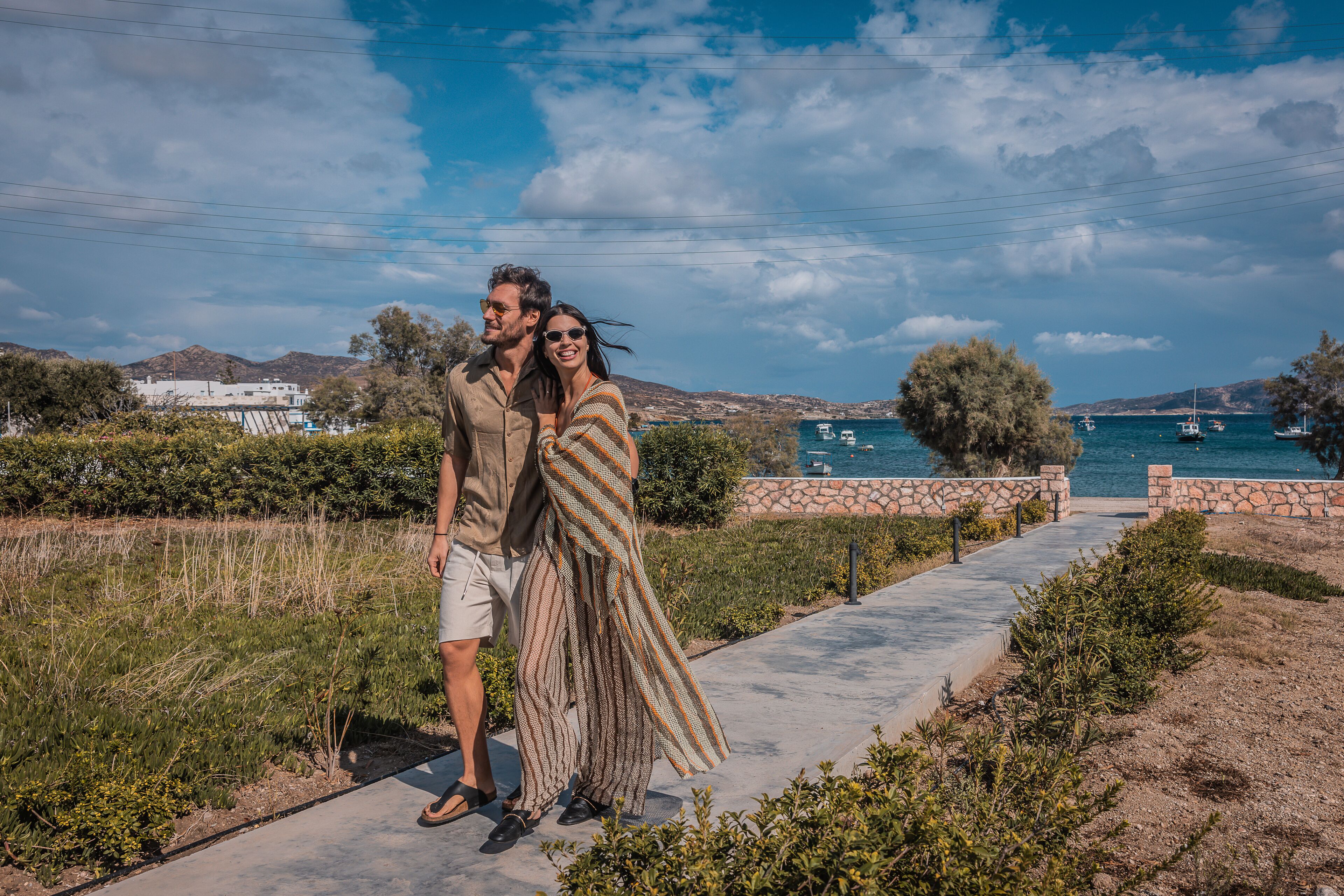 A couple standing on a concrete path with a coastal view, including boats and distant buildings.