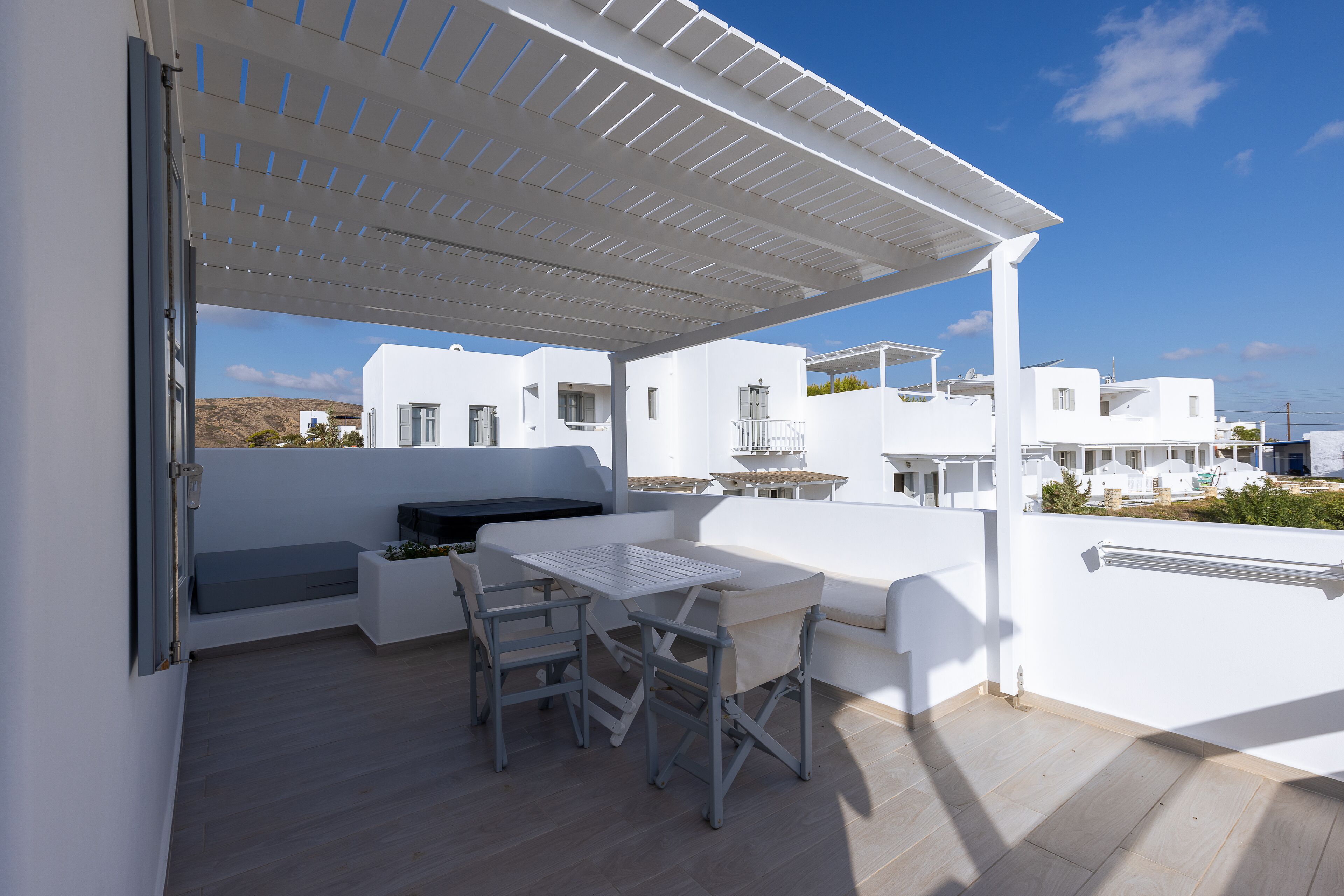 A covered outdoor patio with a table and chairs, white walls, and a view of white buildings in the background.