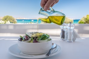 A bowl of salad with olive oil being poured over it, set on a table with a view of the sea and a white tablecloth. - En Milo (Milos)