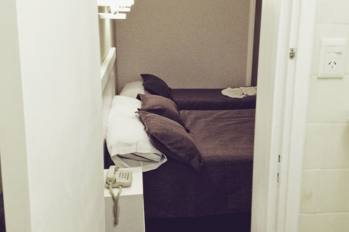 A narrow hallway leading to a bedroom with a bed, pillows, a bedside table, and a telephone.