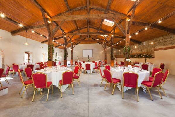 Ein großer Saal mit runden Tischen und roten Stühlen, Holzbalken an der Decke und einer Steinwand im Hintergrund.