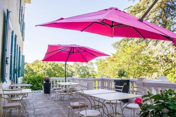 Une terrasse extérieure avec des tables et des chaises, deux grands parasols roses et une balustrade en pierre.