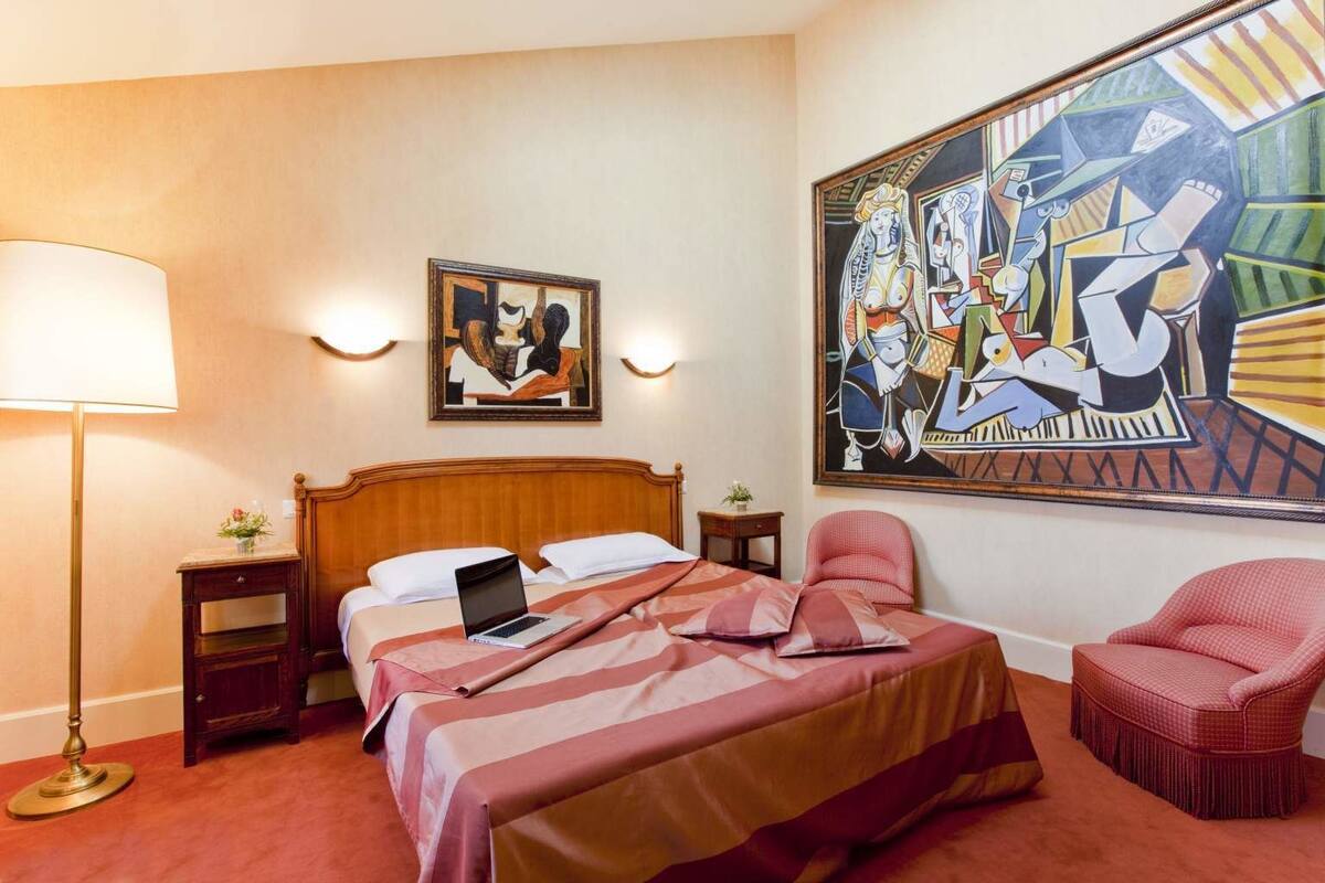 Une chambre d’hôtel avec un lit en bois, un ordinateur portable posé sur le lit, un fauteuil rouge, un tableau accroché au mur et une lampe.