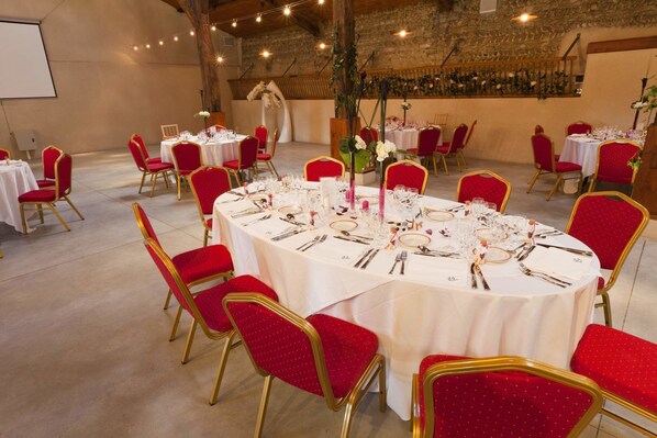 Une salle de banquet avec des tables rondes dressées pour un événement formel, agrémentée de chaises à revêtement rouge et de nappes blanches.