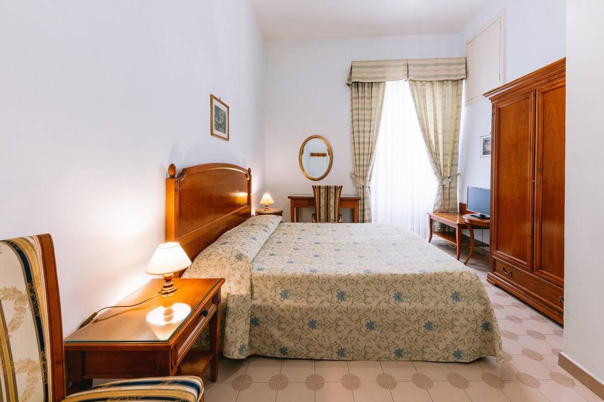 A bedroom with a wooden bed, a bedside table with a lamp, a chair, a desk, a wardrobe, and a window with curtains.