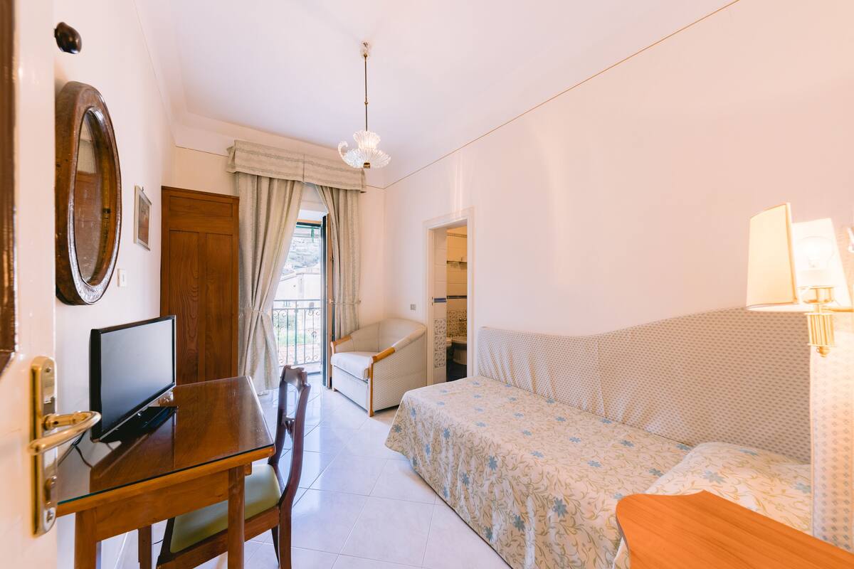 A hotel room with a bed, a desk with a computer, a chair, a window with curtains, and a chandelier.