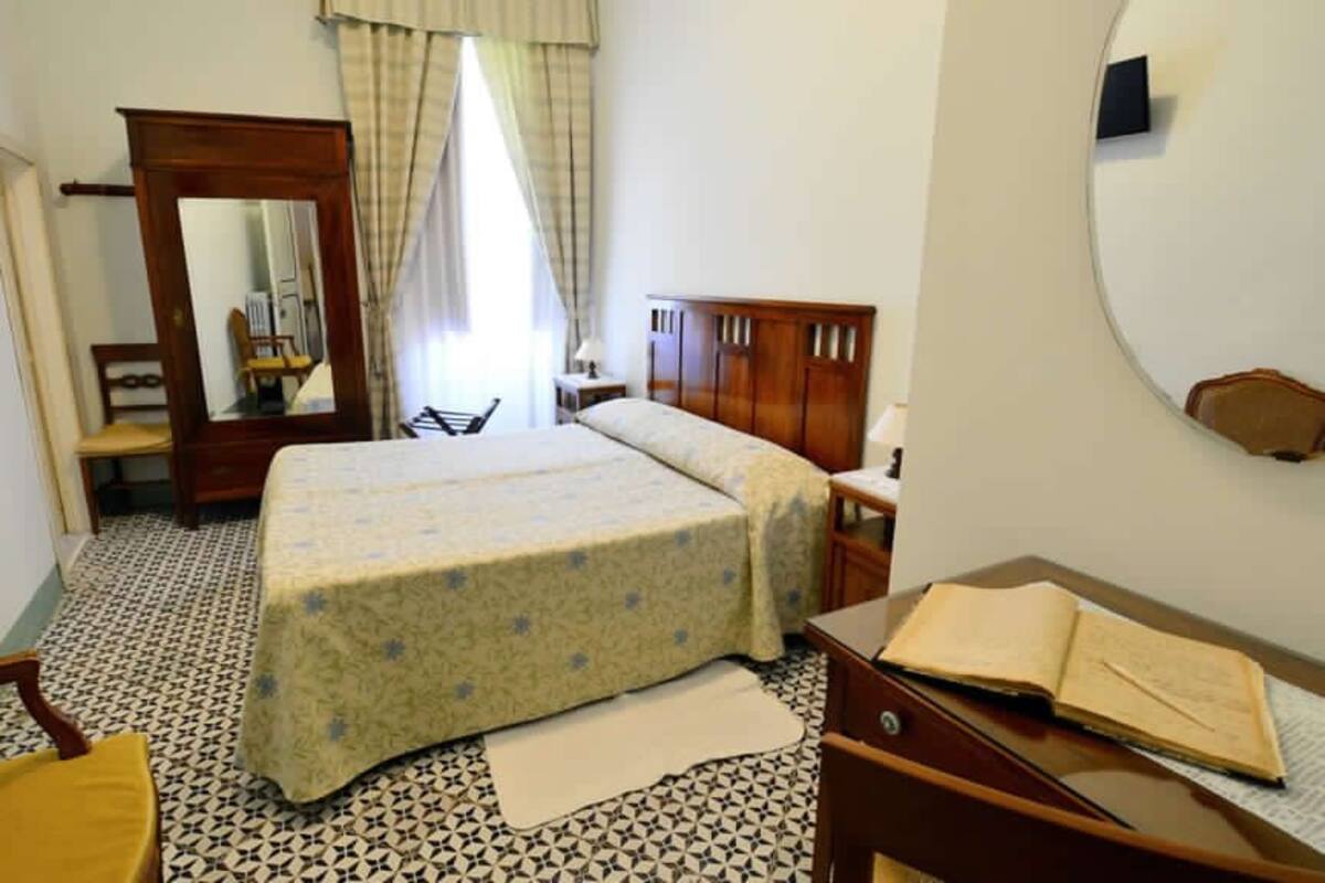 A bedroom with a bed, a wooden wardrobe, a desk with a chair, a mirror, and a window with curtains.