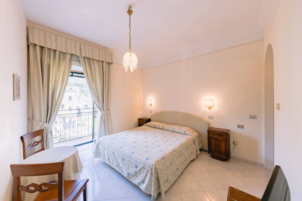 A bedroom with a bed, bedside tables, a chair, a table, and a balcony with curtains.