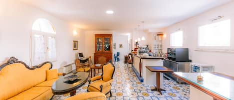 Un salon comprenant un canapé, des fauteuils, une table basse et un téléviseur. Il y a également un coin cuisine avec un réfrigérateur, une cuisinière et des placards. Le sol est recouvert de carreaux décorés.