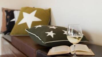 Un coin lecture confortable avec un canapé, un coussin orné de motifs en forme d’étoiles, un livre ouvert et un verre de vin blanc posé sur une table en bois.