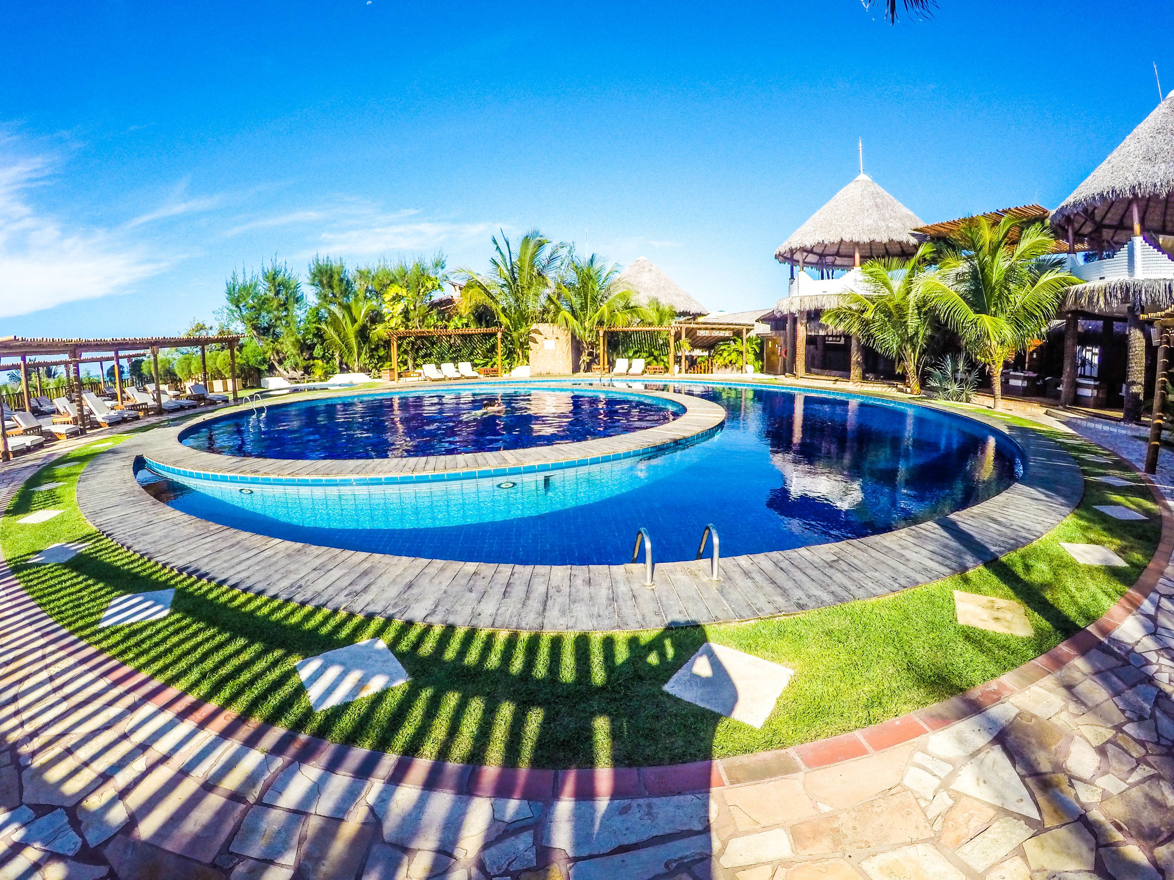 Una piscina circular con un muelle de madera, palmeras y estructuras con techo de paja.