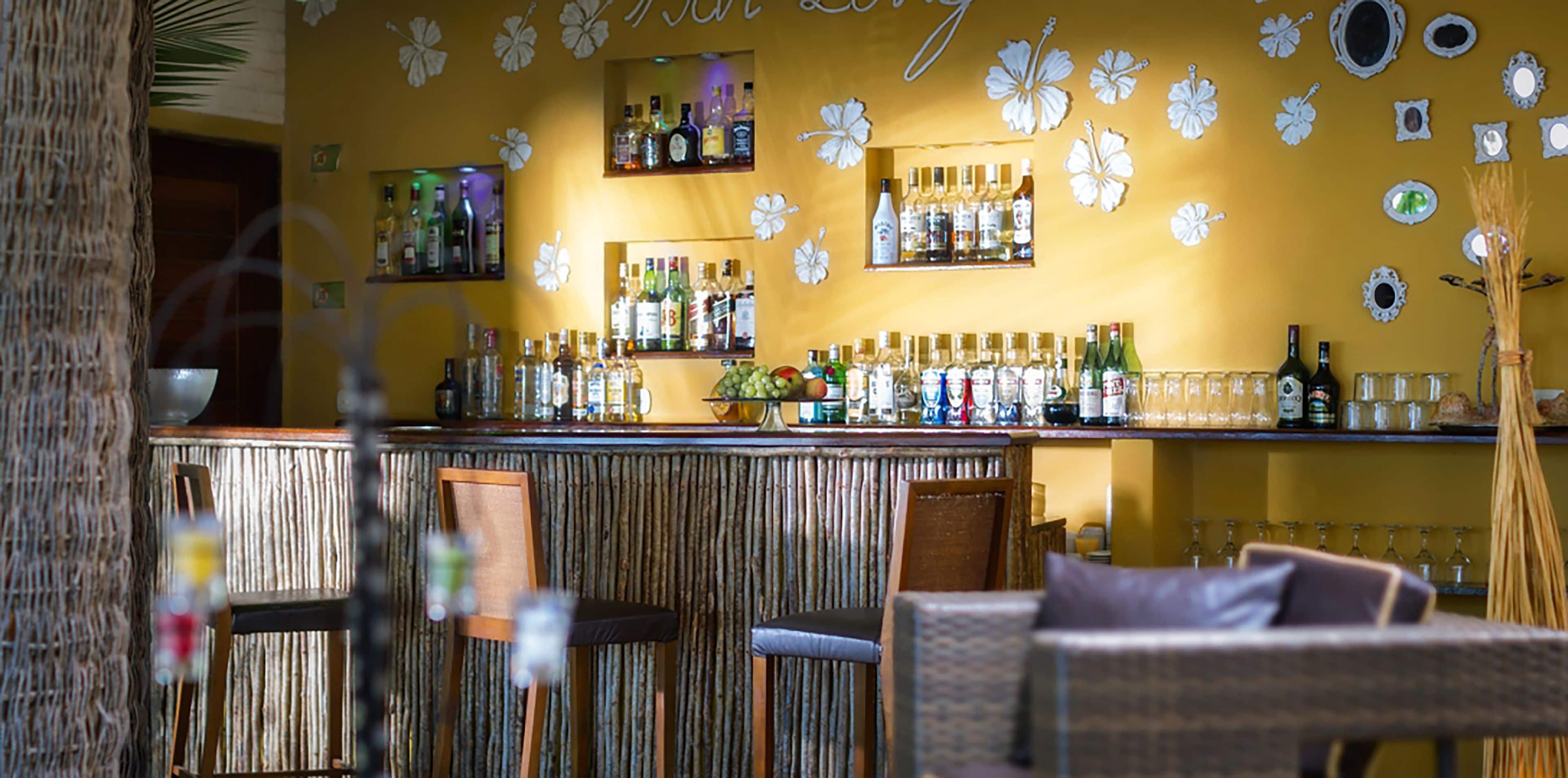 A bar area with a wooden bar counter, shelves with bottles, and a wall decorated with white floral patterns.
