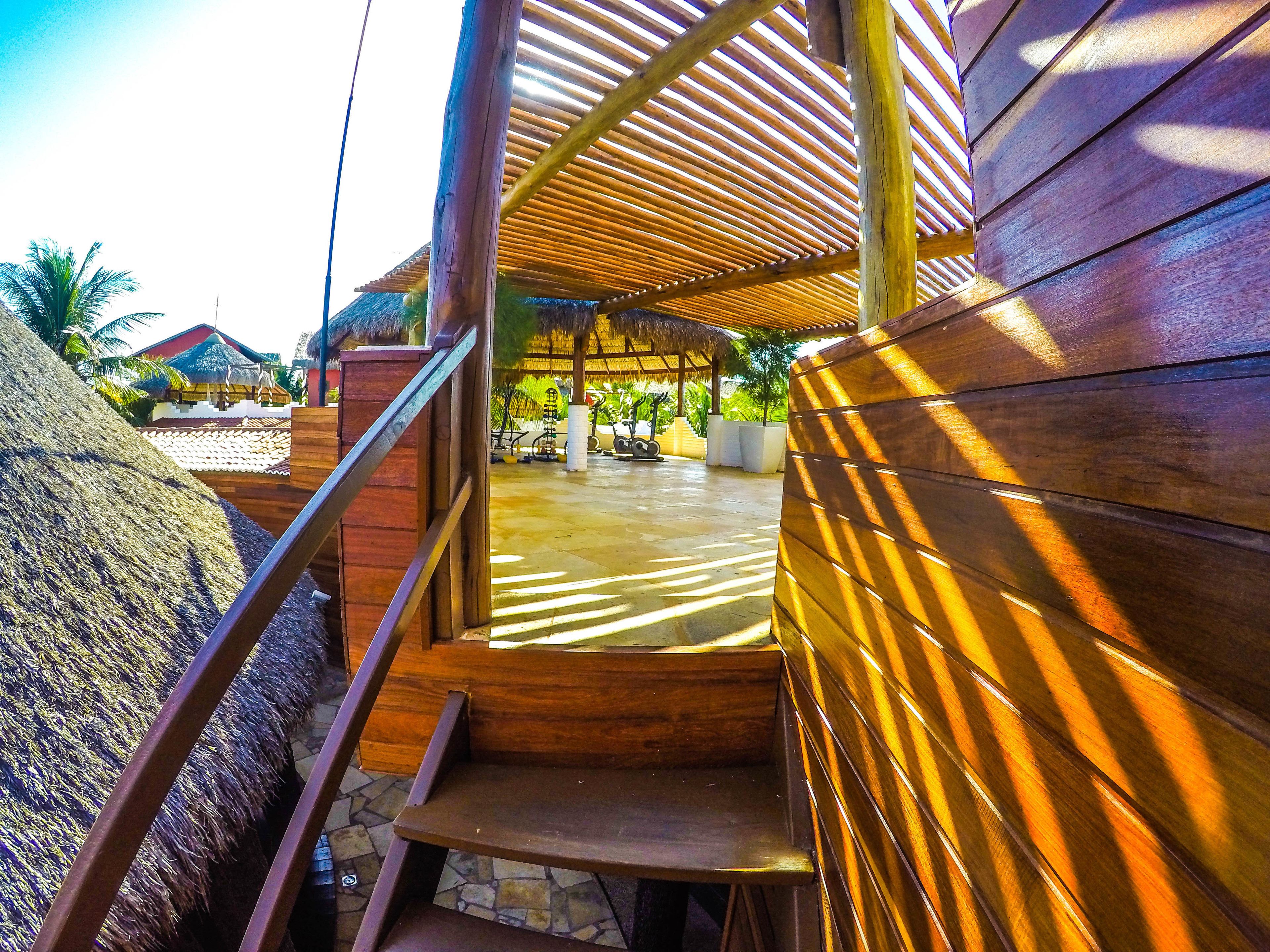 A wooden staircase leading to a covered outdoor area with a thatched roof, stone flooring, and a view of a tropical resort setting.