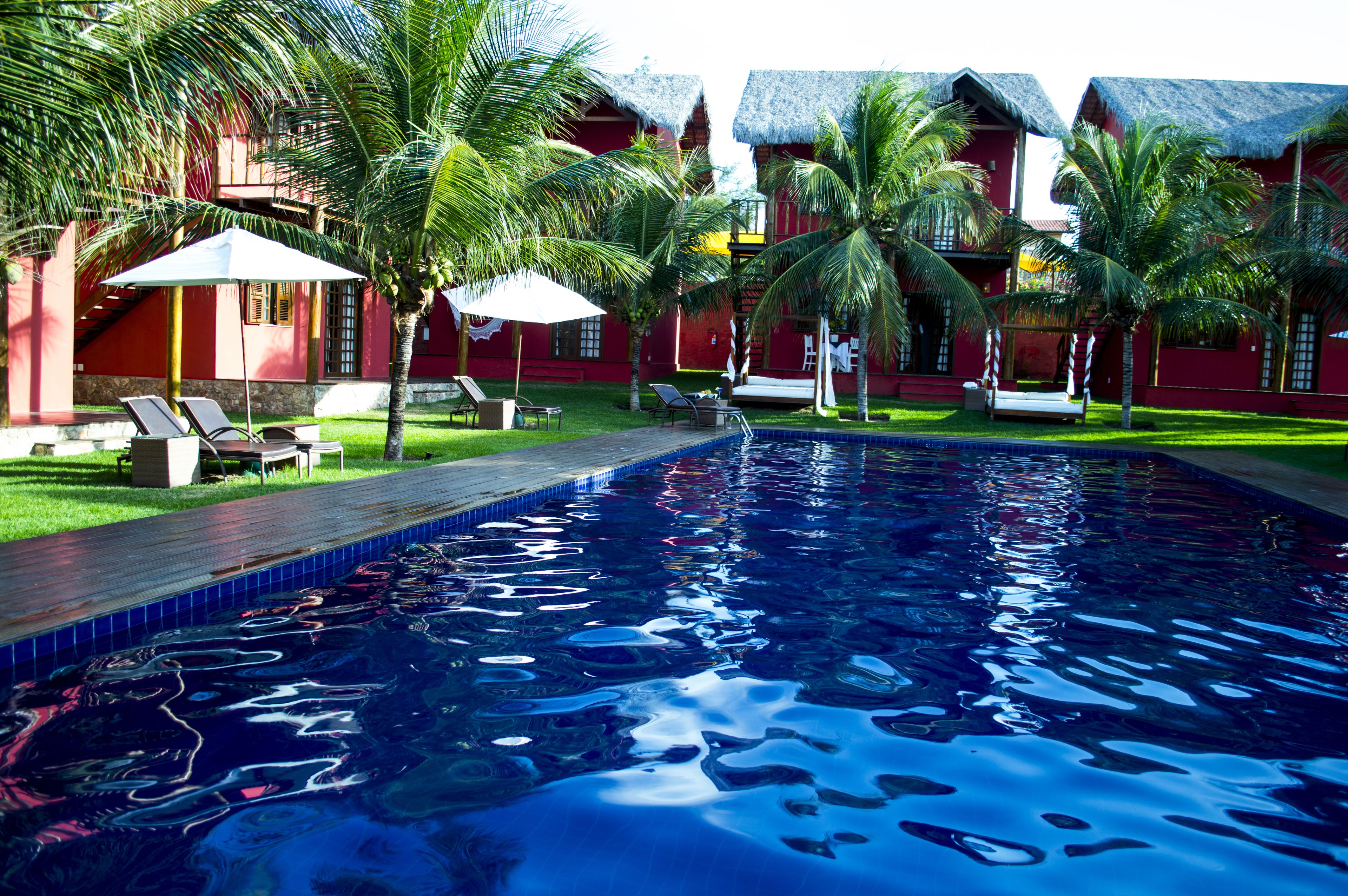Una piscina con sillas de descanso y sombrillas, rodeada de palmeras y edificios rojos con techos de paja.