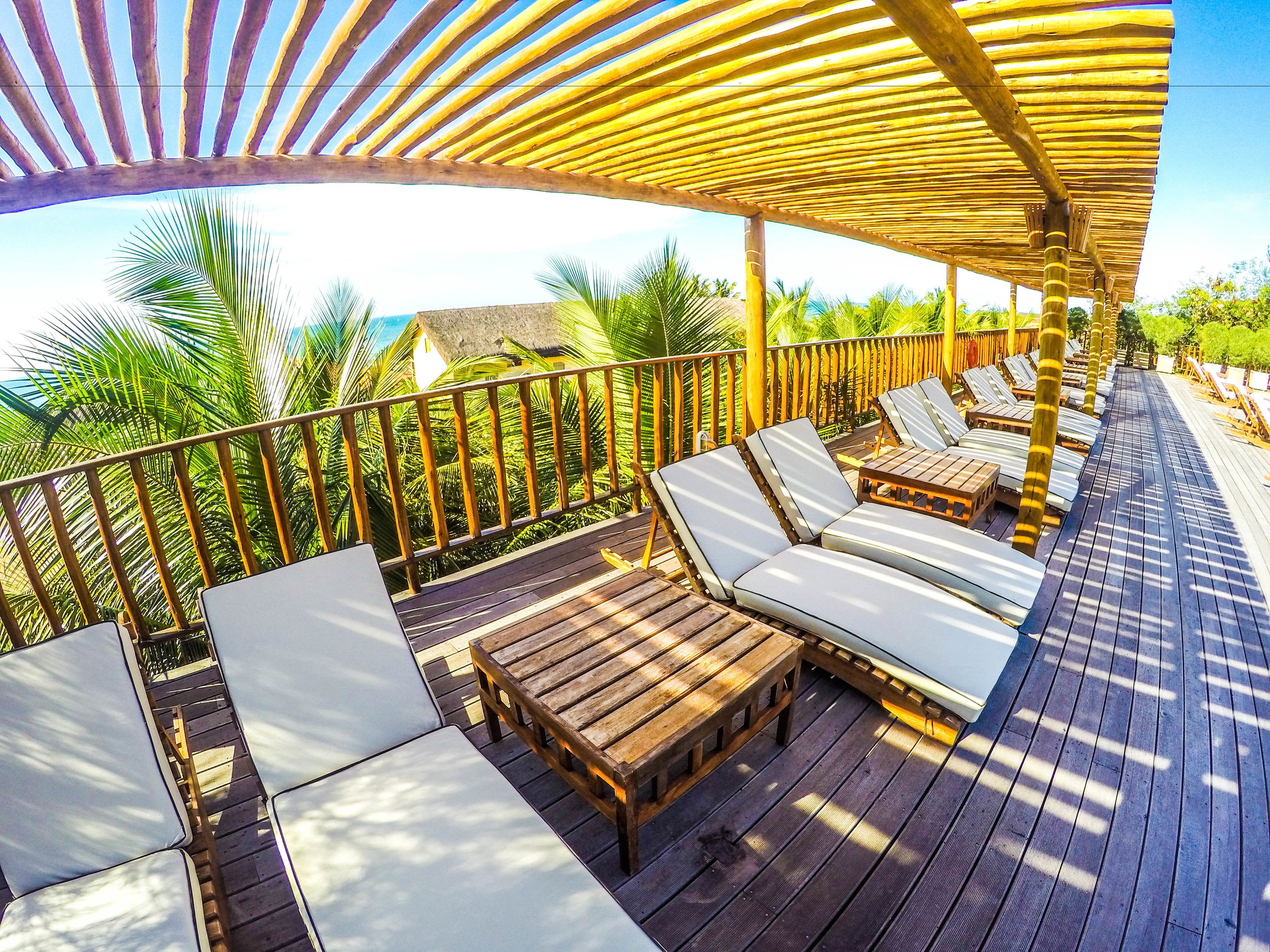 Una terraza de madera con sillones y una mesa, rodeada de palmeras y un cielo despejado.