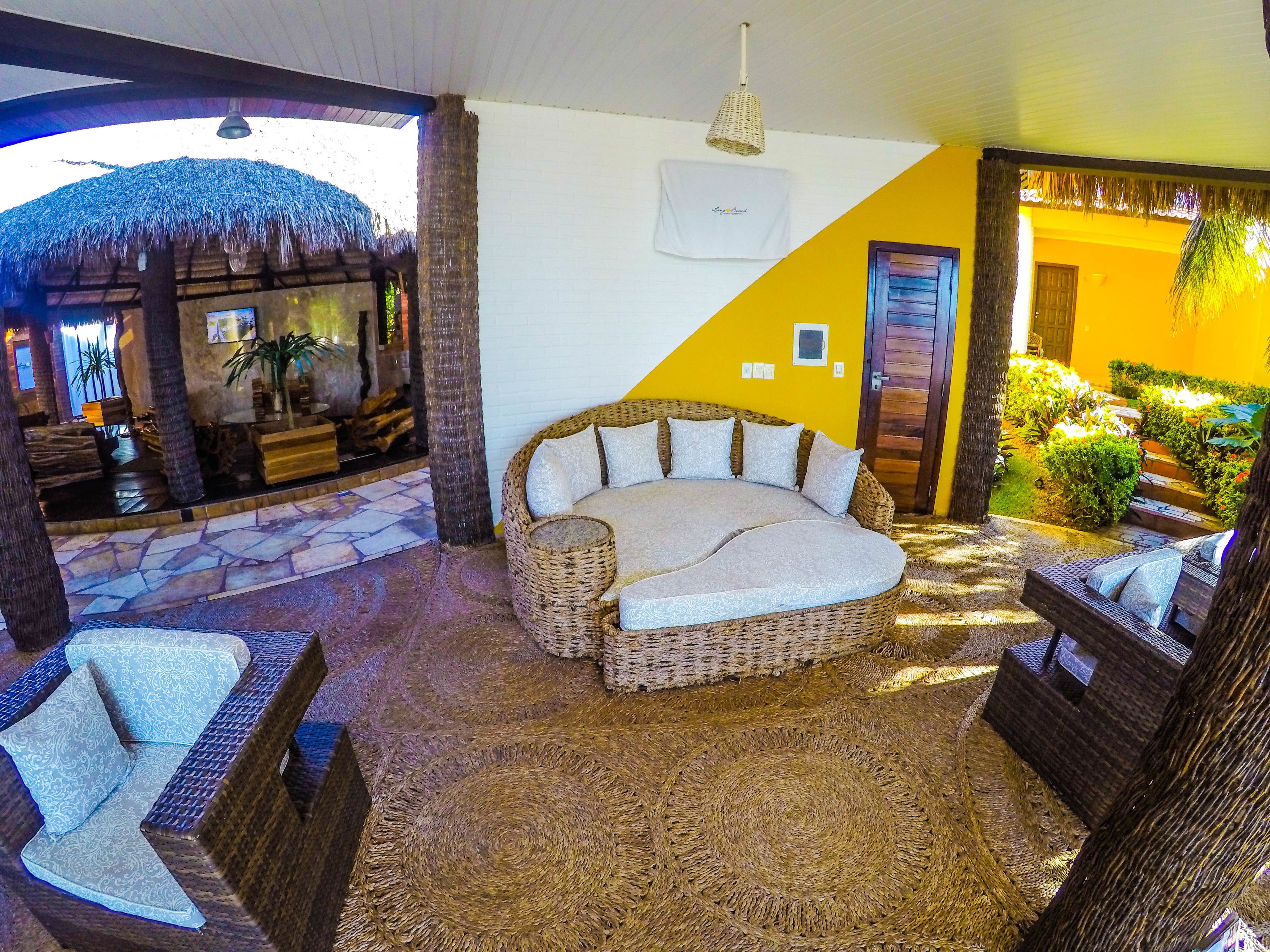 A cozy lounge area with a wicker sofa, a patterned armchair, and a small round table. A thatched roof structure and a stone wall are visible in the background.