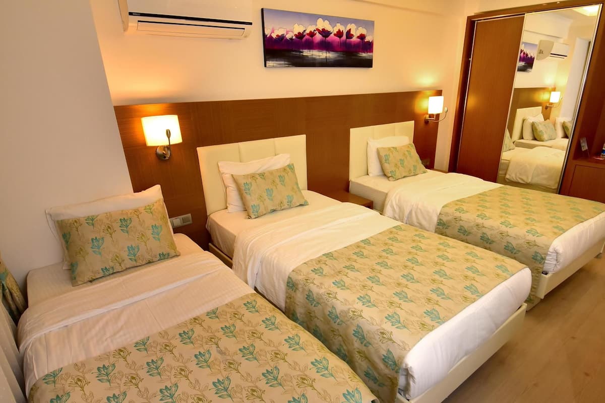 A hotel room with three beds, each with floral-patterned bedding, a ceiling light, an air conditioning unit, and a wall-mounted painting.