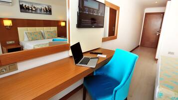 Habitación de hotel con un escritorio de madera, una computadora portátil, una silla azul, una cama con ropa de cama floral, un televisor montado en la pared y una pintura de un paisaje urbano.