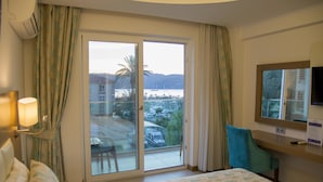 Habitación de hotel con cama, escritorio, silla, televisor y balcón con vistas al mar y edificios.