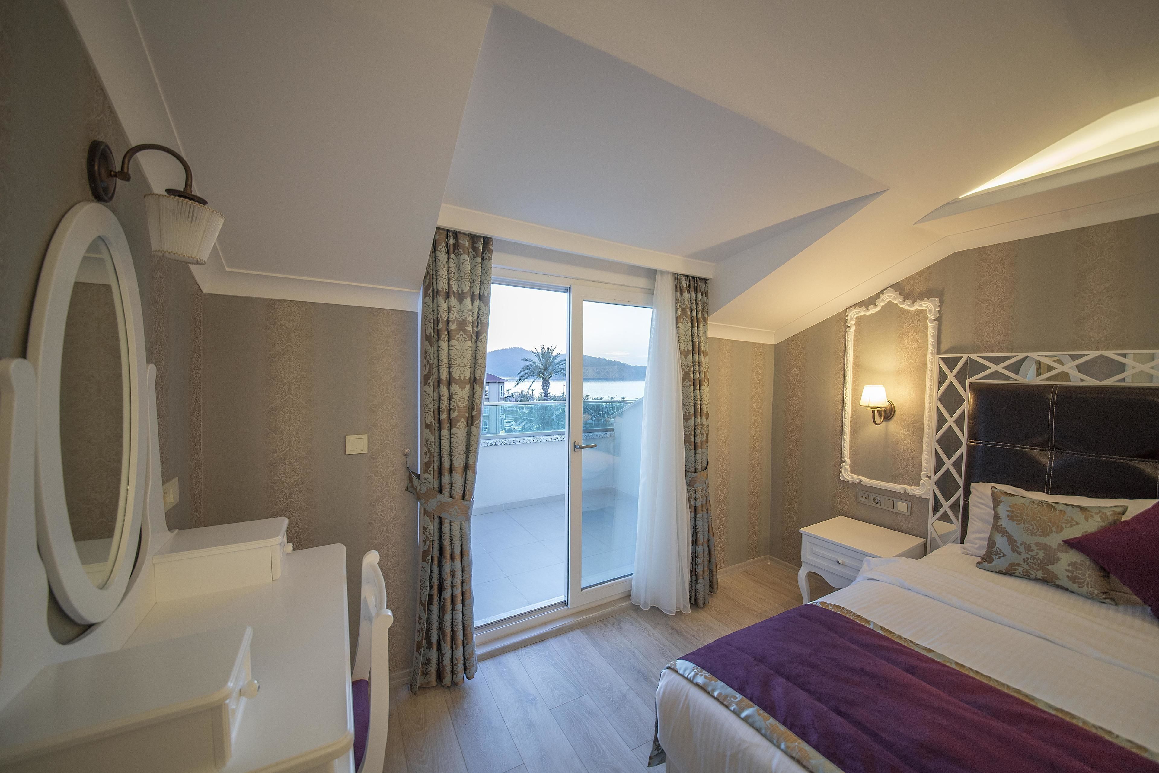 A hotel room with a bed, a nightstand, a dresser, a mirror, and a sliding glass door leading to a balcony with a view of the sea.