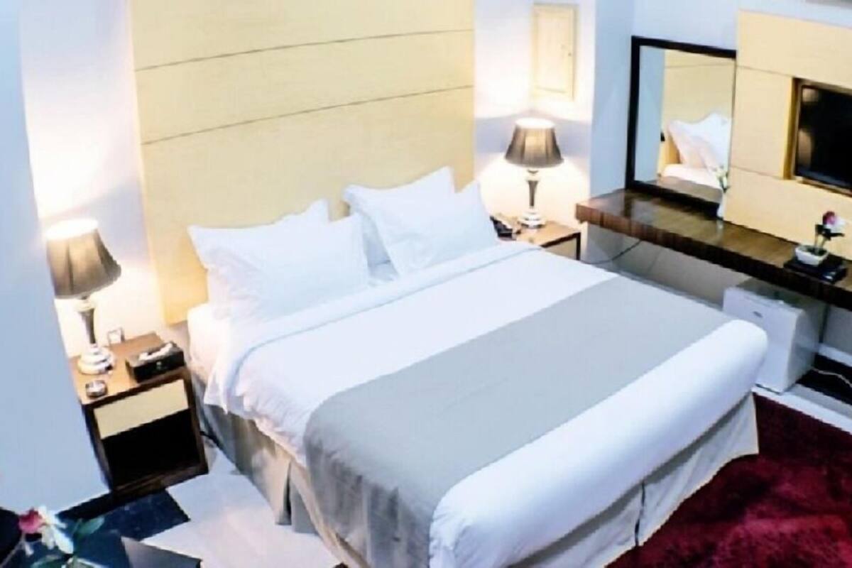 A neatly made bed with white linens, a bedside table with a lamp, a dresser with a mirror, and a TV on the dresser.