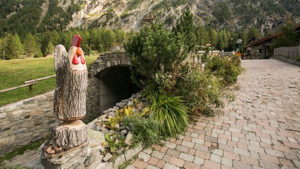 A rustic stone structure with a wooden sculpture of a rooster, a stone bridge, and a mountainous backdrop. - La Barme (Cogne)