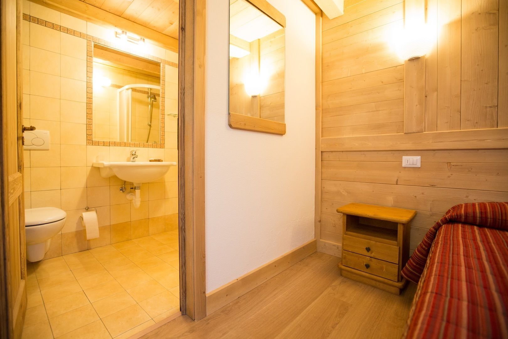 A small, cozy room with a wooden floor, a single bed with a striped blanket, a wooden nightstand, a small drawer, a white sink, a toilet, and a mirror.