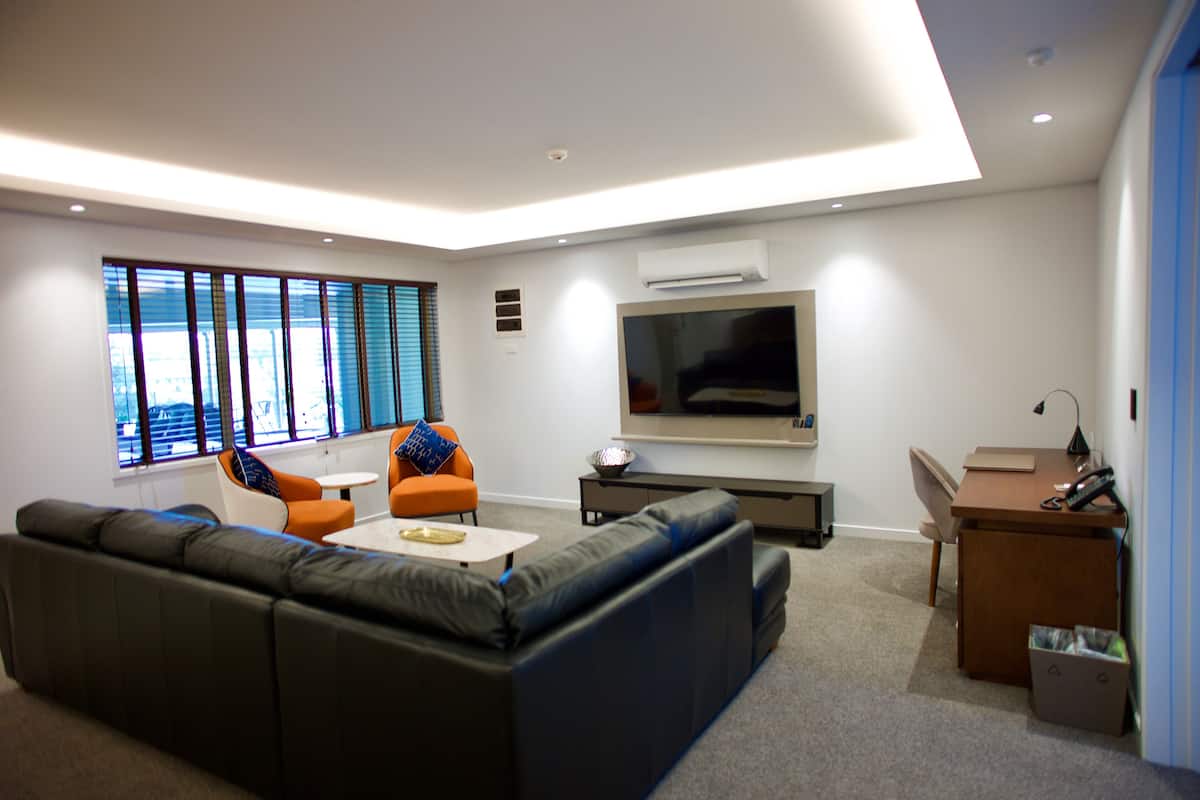Un salon moderne avec un canapé d’angle, une table basse et deux fauteuils. Un téléviseur à écran plat est fixé au mur, il y a un bureau avec une chaise et une corbeille à déchets.