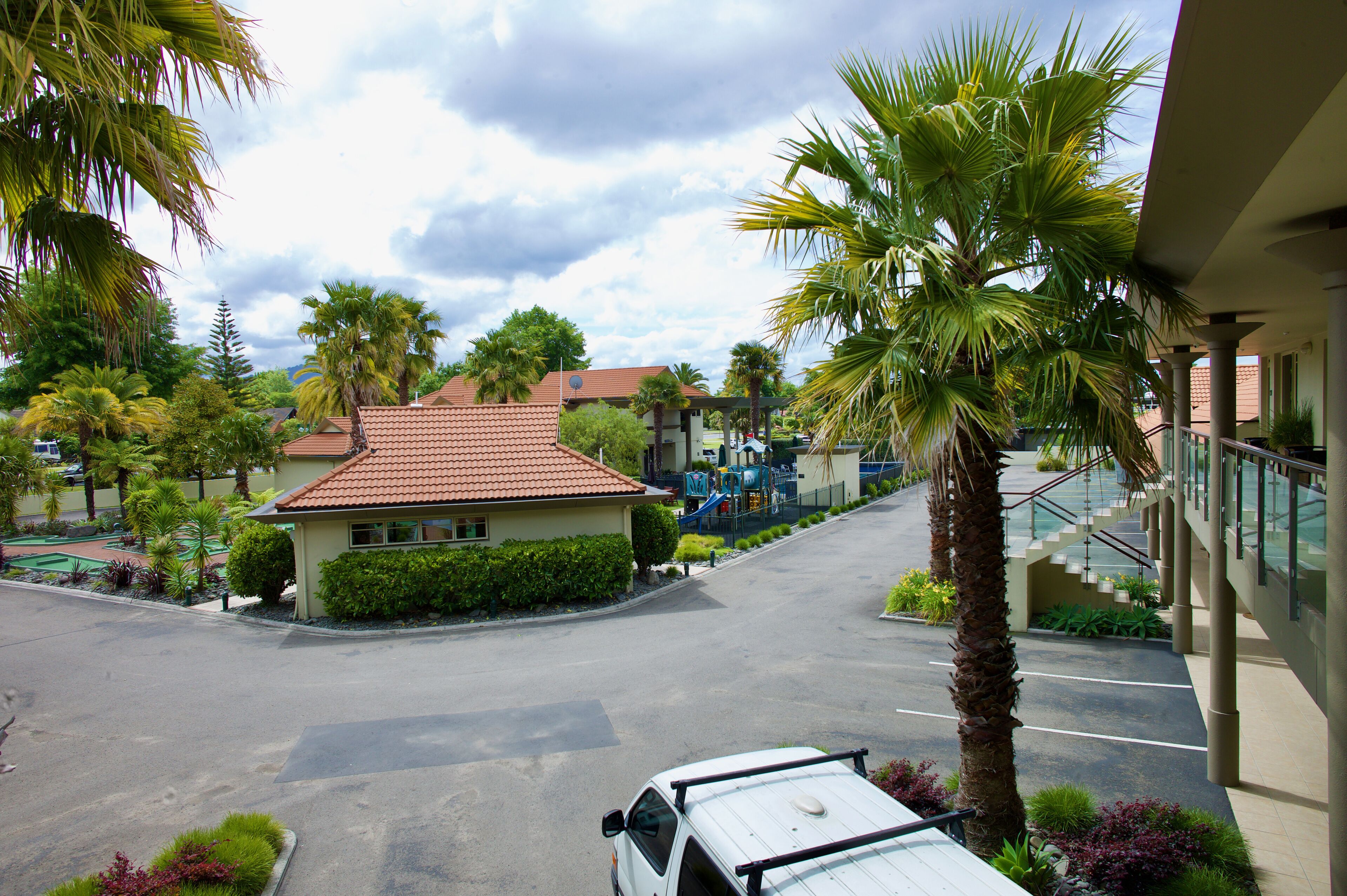 A parking area with a white car, a building with a red roof, and palm trees.