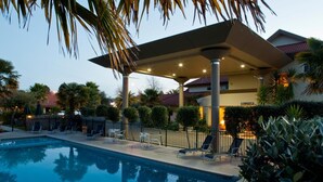 A swimming pool with lounge chairs and a covered area, surrounded by palm trees and a building with a sign. - Regal Palms Resort (Rotorua)