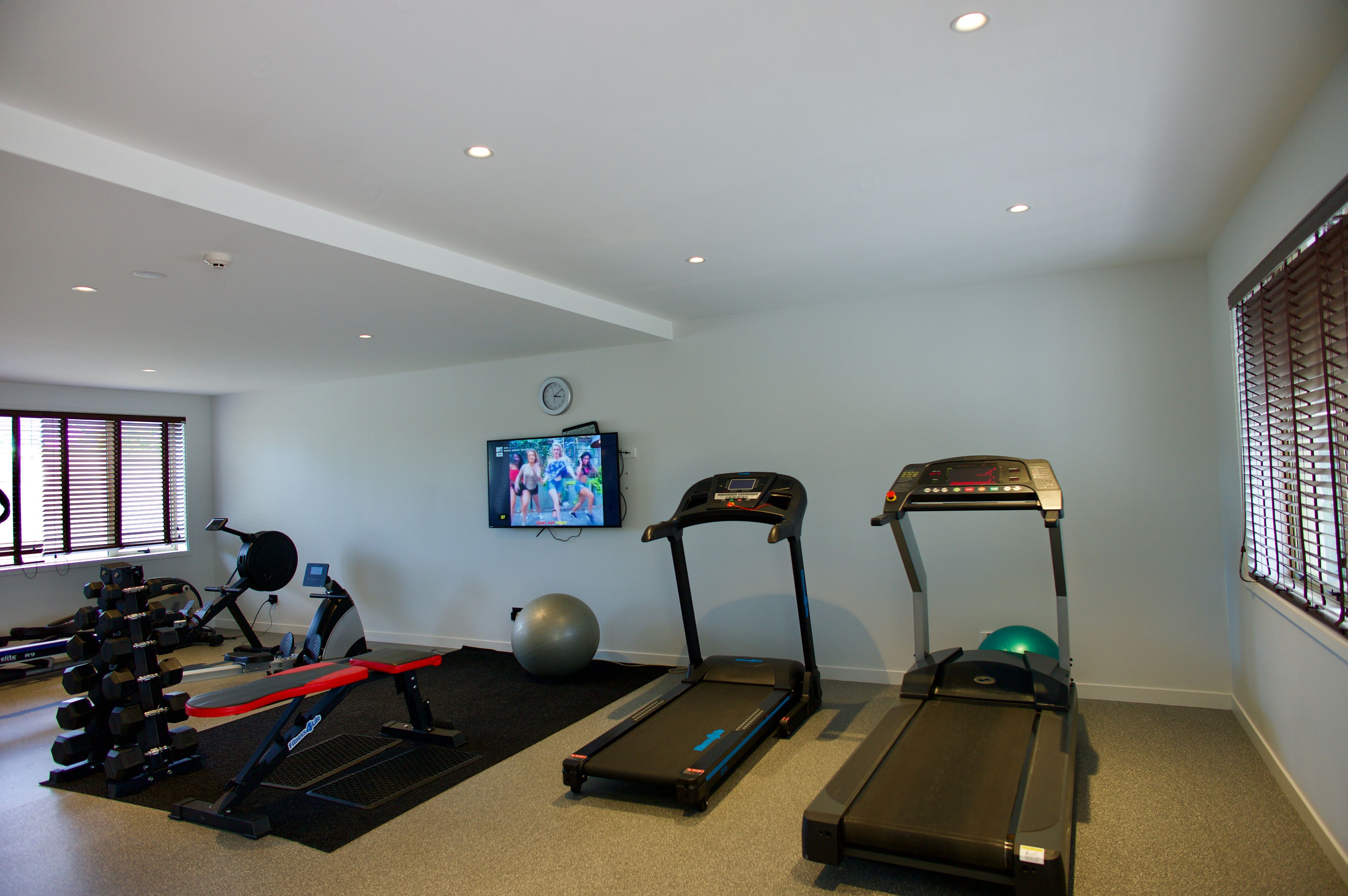 Een sportschool thuis met loopbanden, gewichten, een hometrainer en een televisie waarop een trainingsvideo wordt afgespeeld.