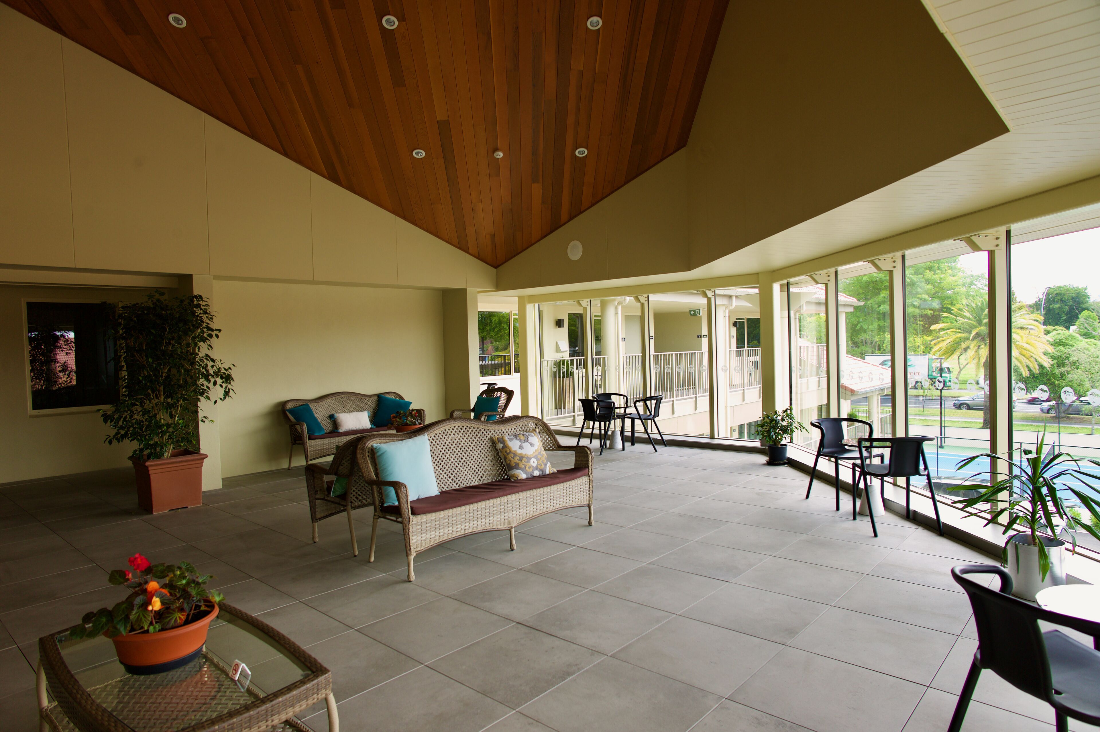 A spacious indoor area with a high ceiling, a wicker sofa with cushions, a glass table with a potted plant, and several chairs.
