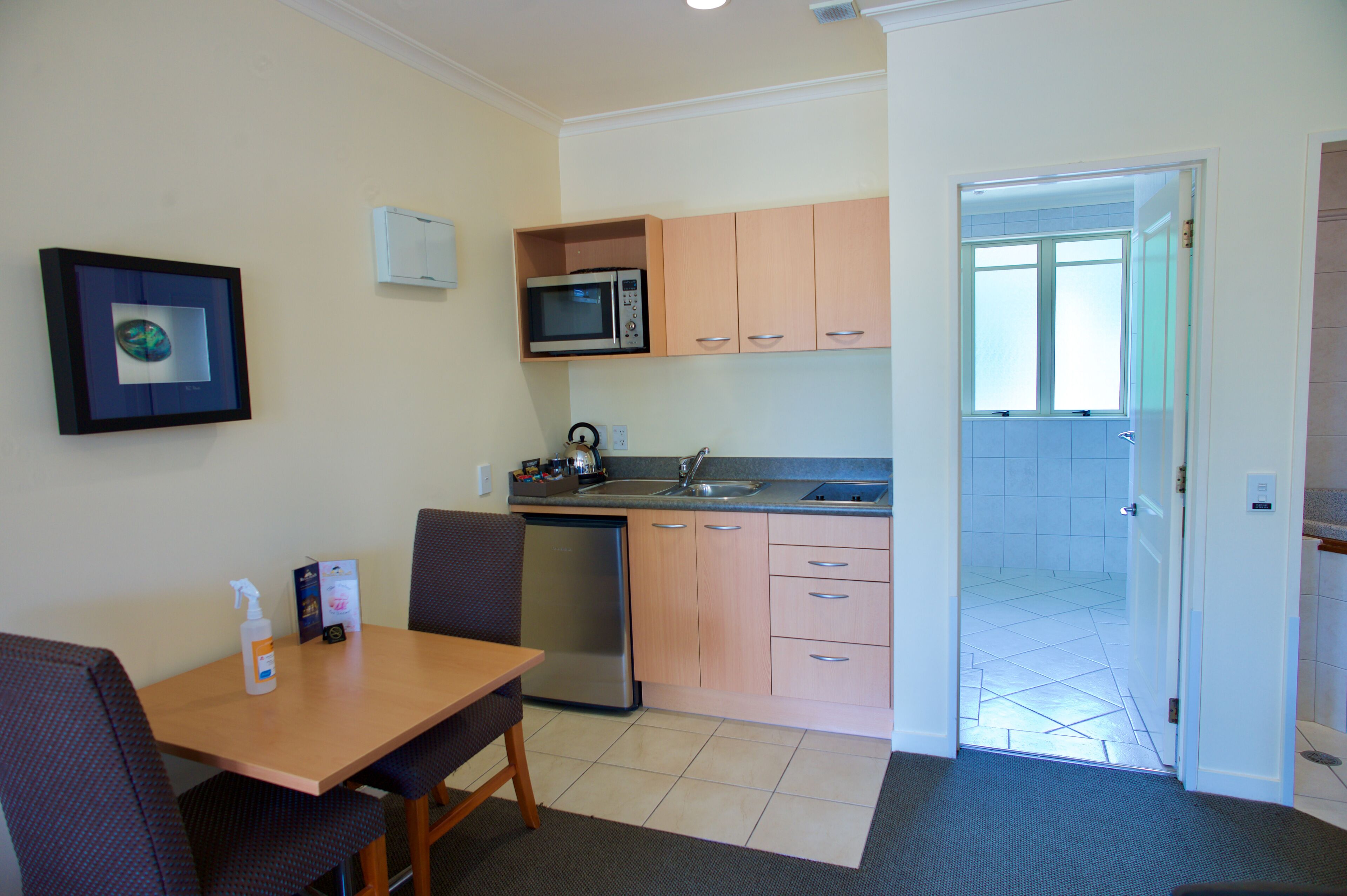 A compact kitchen with a microwave, sink, and dishwasher. There is a dining table with chairs and a framed picture on the wall.