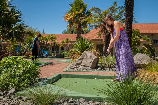 Un campo de mini golf con superficie de césped verde, rodeado de palmeras y rocas.