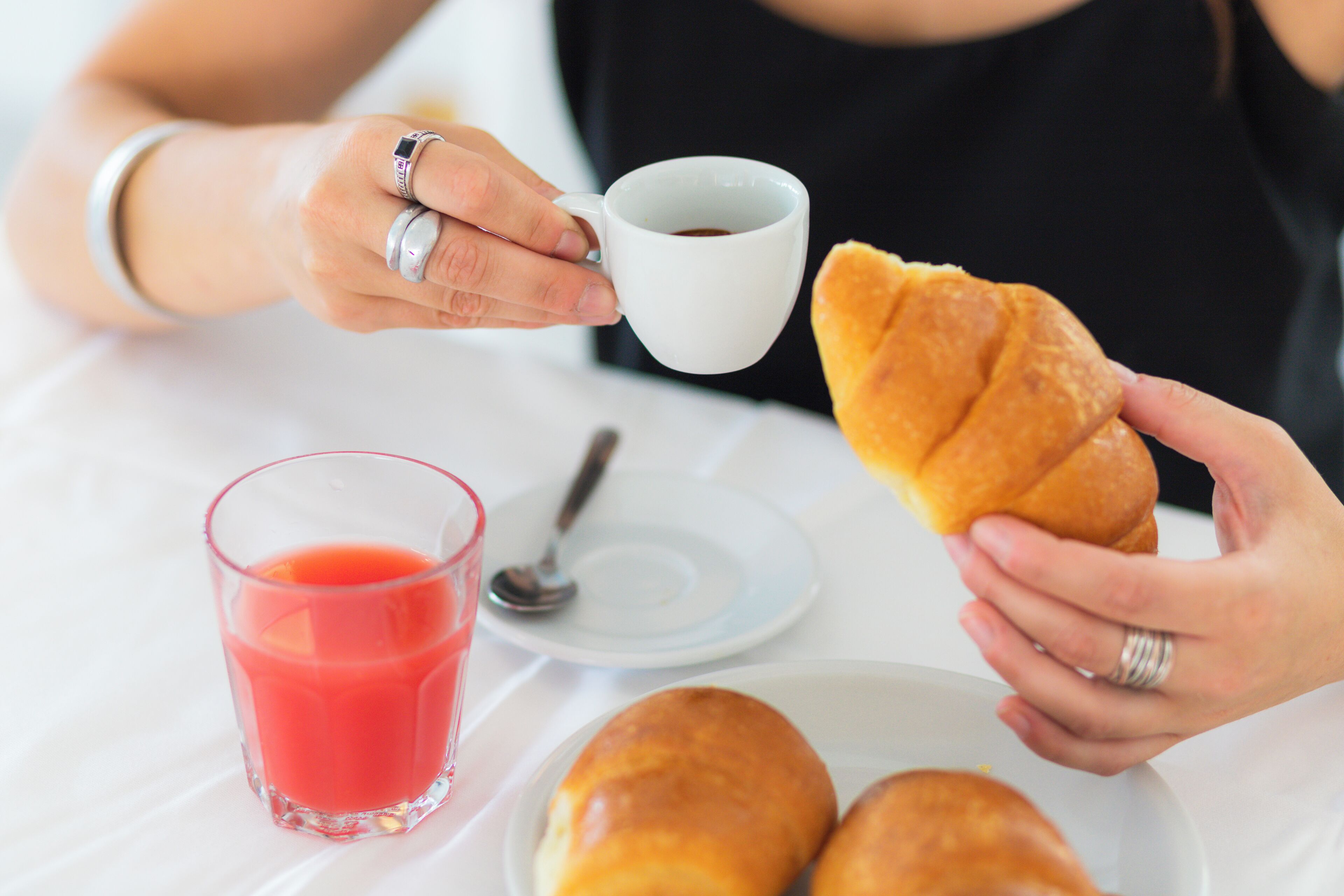 Una persona sosteniendo una taza de café y un croissant, junto con un vaso de jugo y una cuchara sobre una mesa blanca.