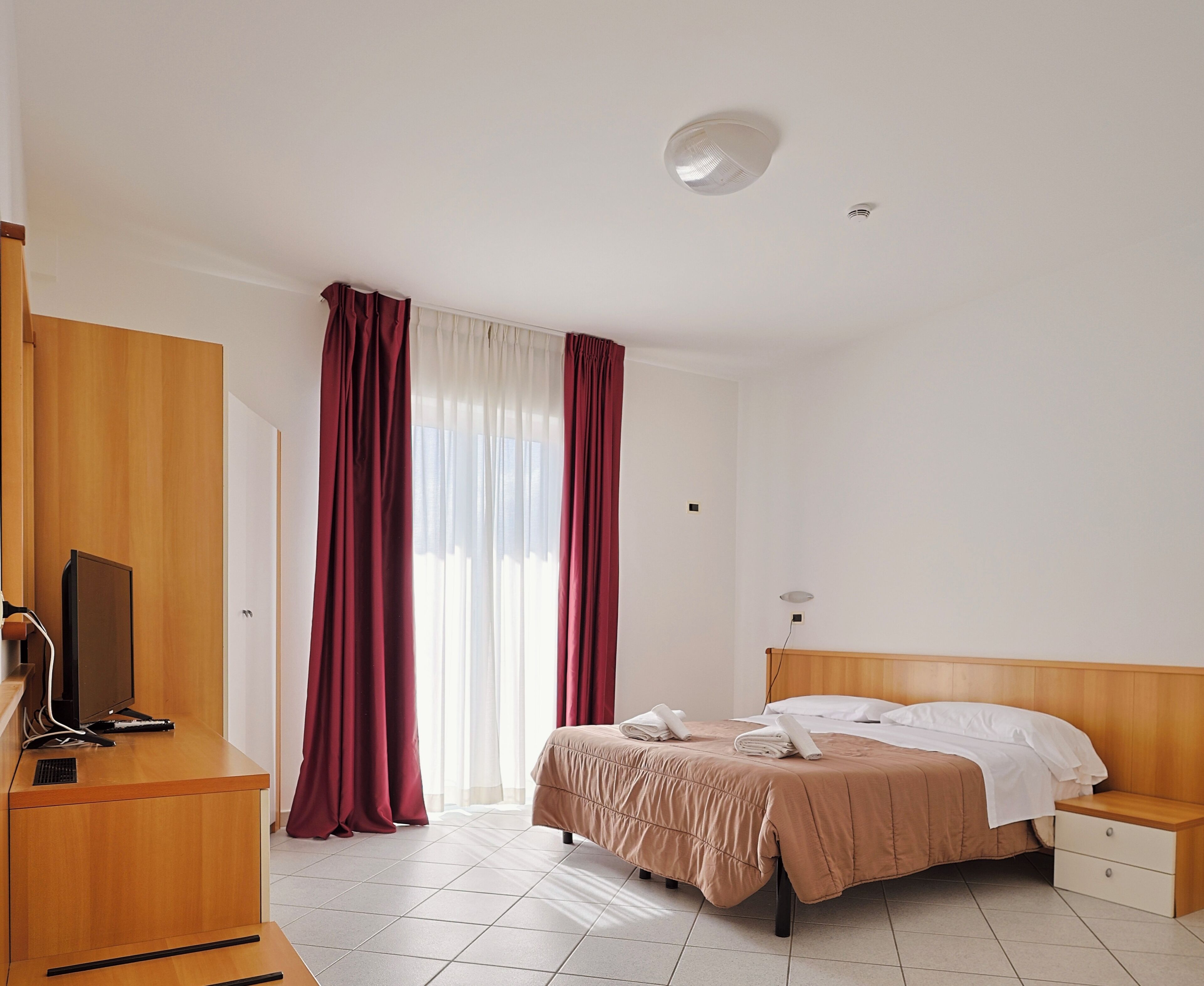 A hotel room with a bed, a desk with a computer, a TV, and a window with curtains.
