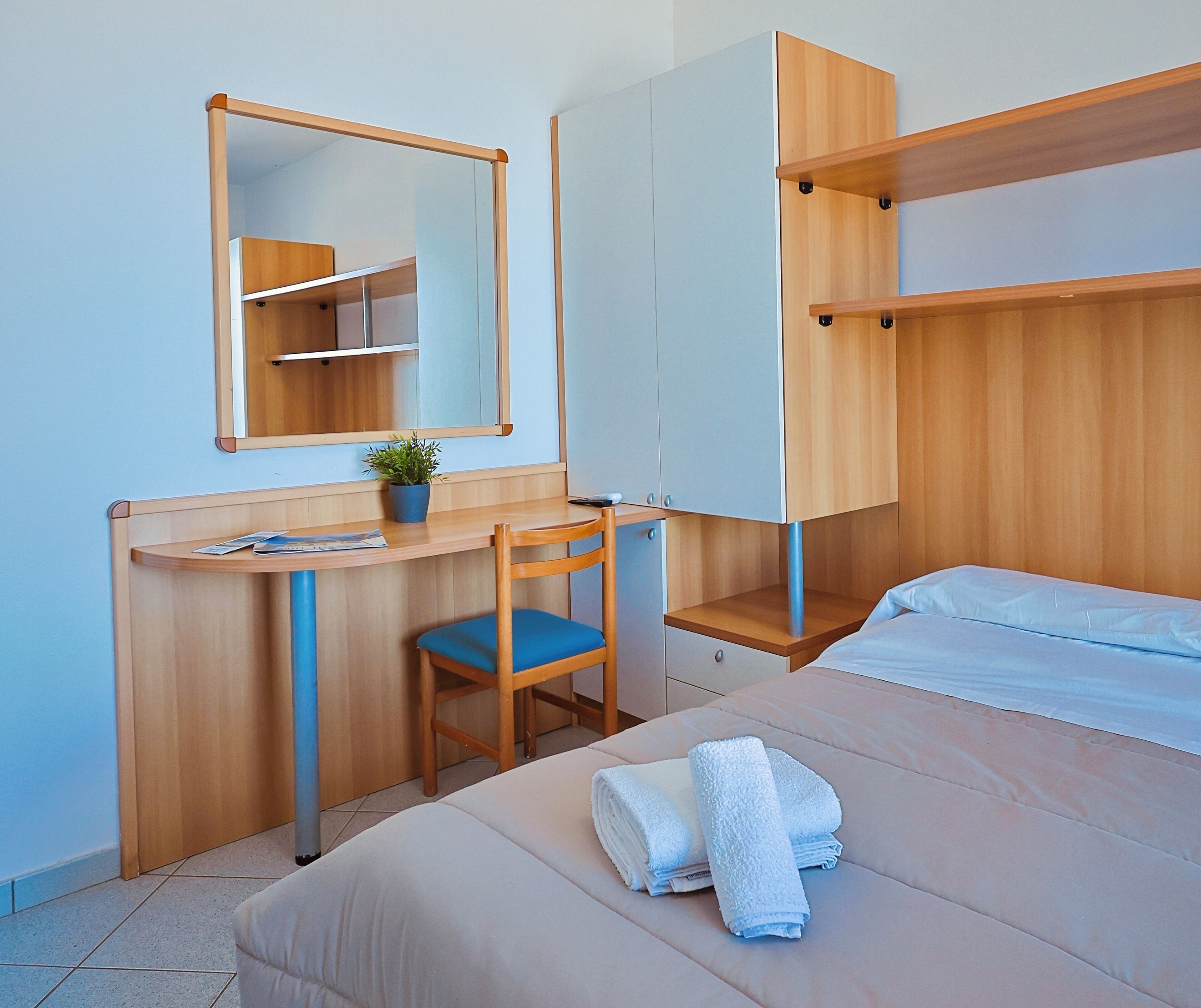 A compact room with a bed, a desk with a chair, a mirror, and a shelf.
