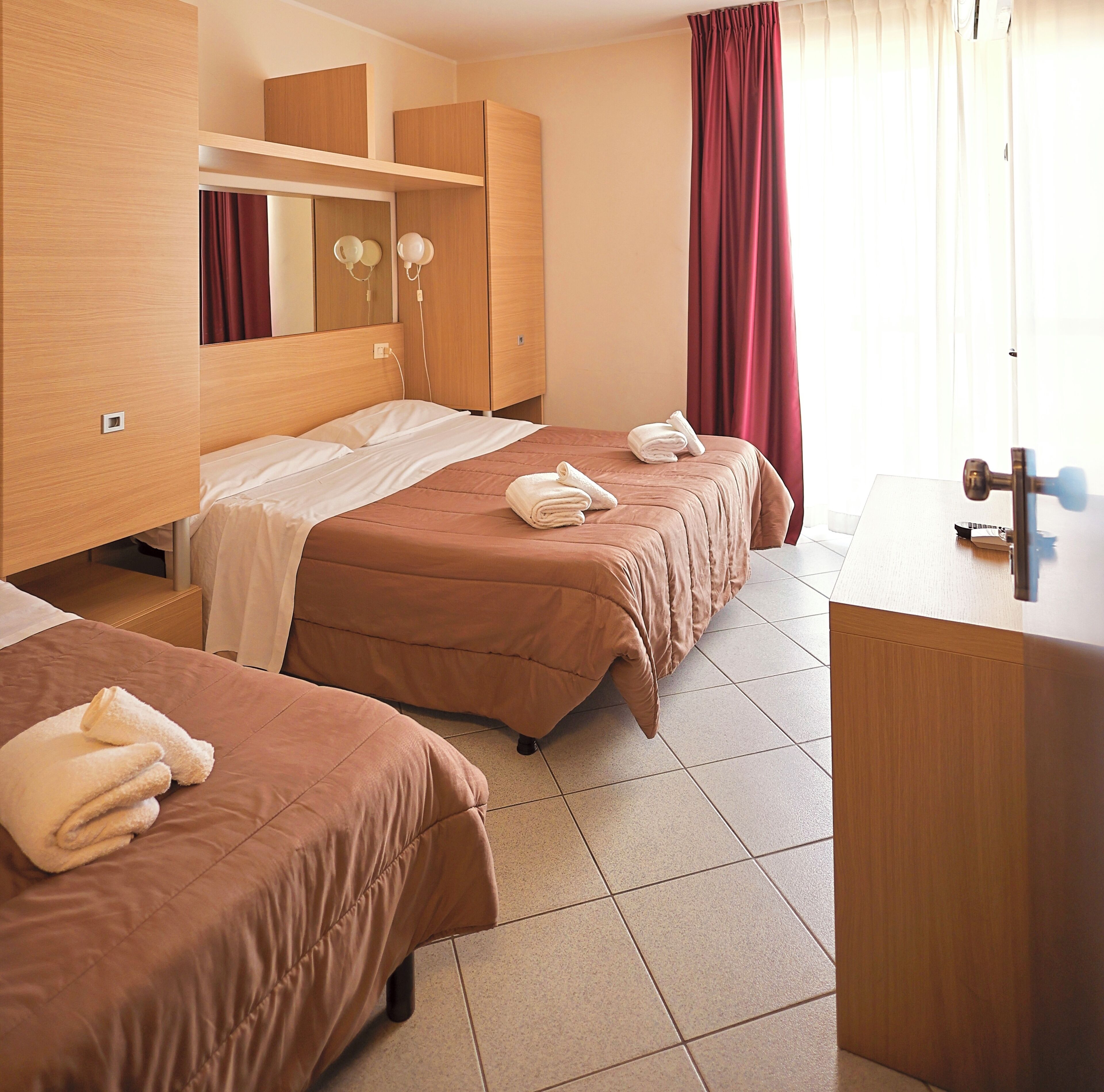 Quarto de hotel com duas camas, um guarda-roupa de madeira, um espelho e uma pequena escrivaninha com espelho.