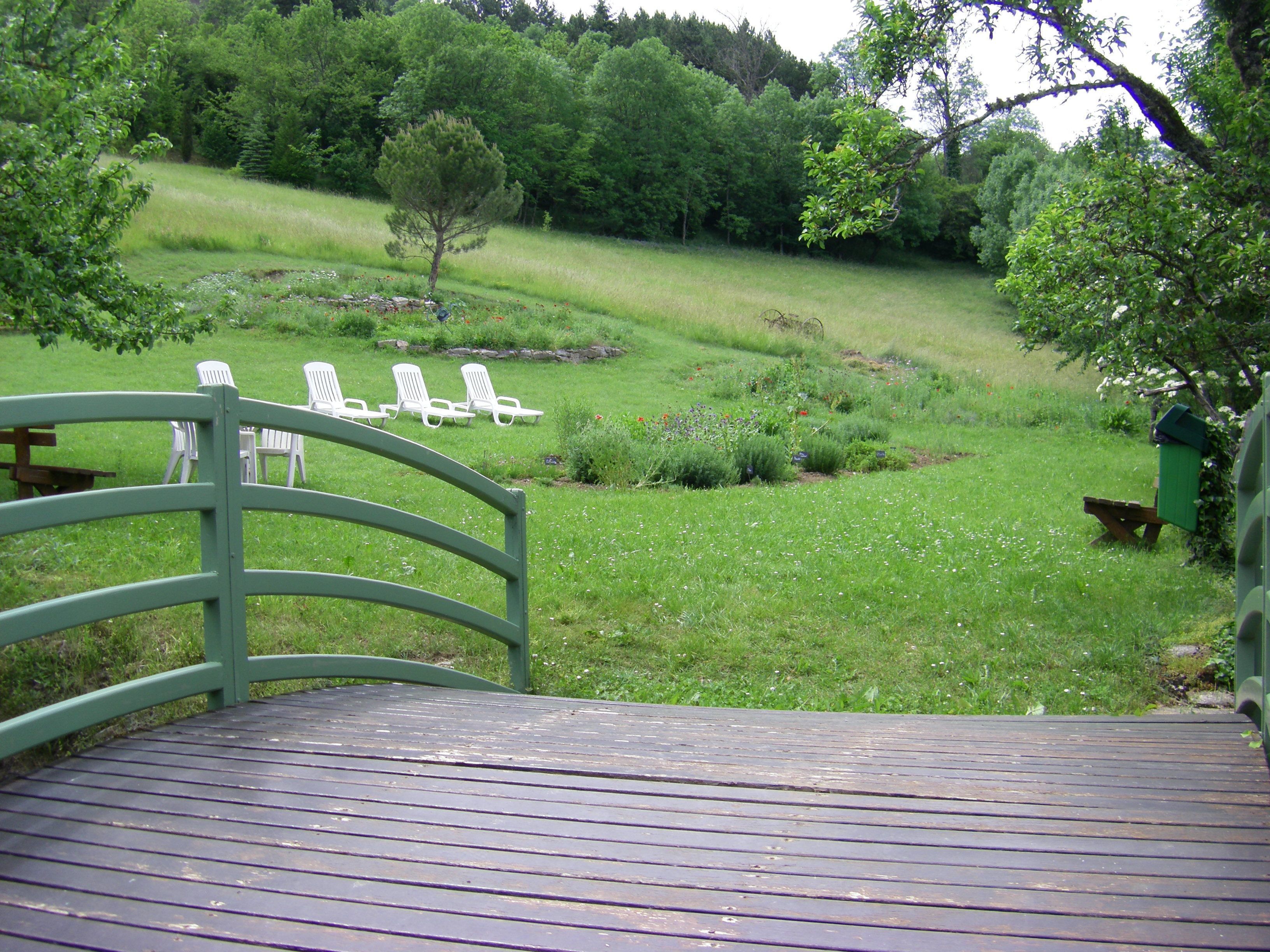 A wooden deck with a curved railing, overlooking a grassy area with white lounge chairs and a small tree.