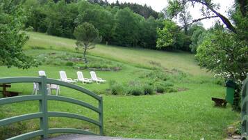 A wooden deck with a curved railing, overlooking a grassy area with white lounge chairs and a small tree.