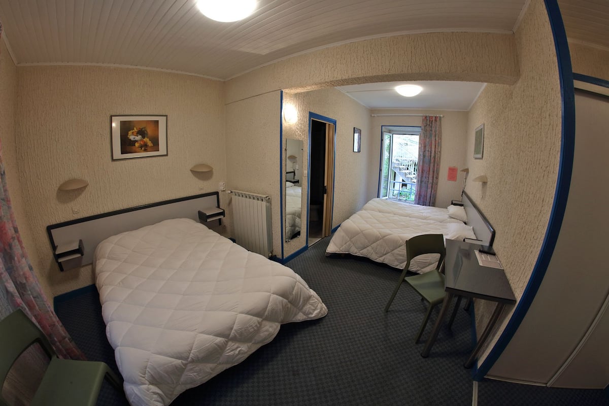 Une chambre d’hôtel avec deux lits, un bureau, une chaise, un radiateur et une fenêtre avec des rideaux.