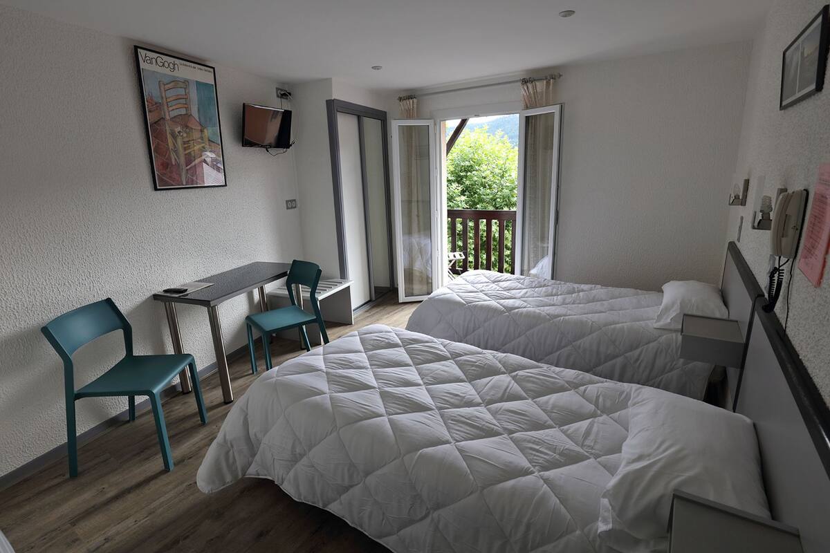 Une chambre avec deux lits, un bureau, des chaises, une télévision et un balcon donnant sur un espace verdoyant.
