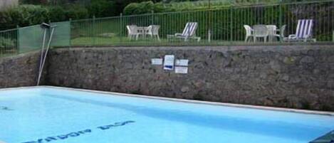 A swimming pool with a stone wall and a metal ladder. There are lounge chairs and umbrellas in the background.