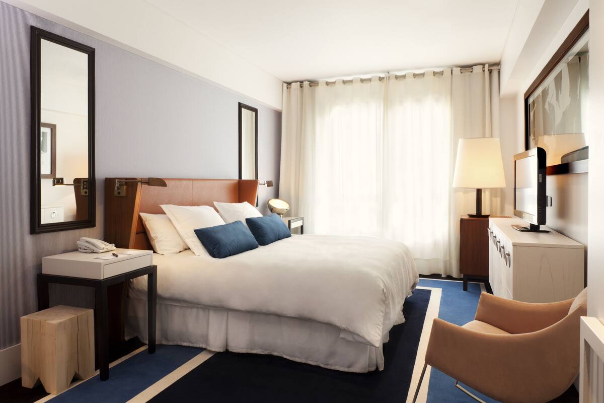 A hotel room with a large bed, bedside tables, a desk, a chair, a mirror, and a window with curtains.