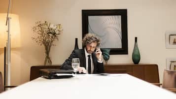 Un hombre con traje sentado a una mesa con una computadora portátil, un vaso de agua y un maletín. Hay una lámpara, un jarrón con flores y una obra de arte enmarcada en la pared detrás de él.