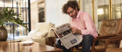 Una persona leyendo un periódico en una sala de estar moderna con una mesa de centro, sillones de cuero y un ventanal grande.