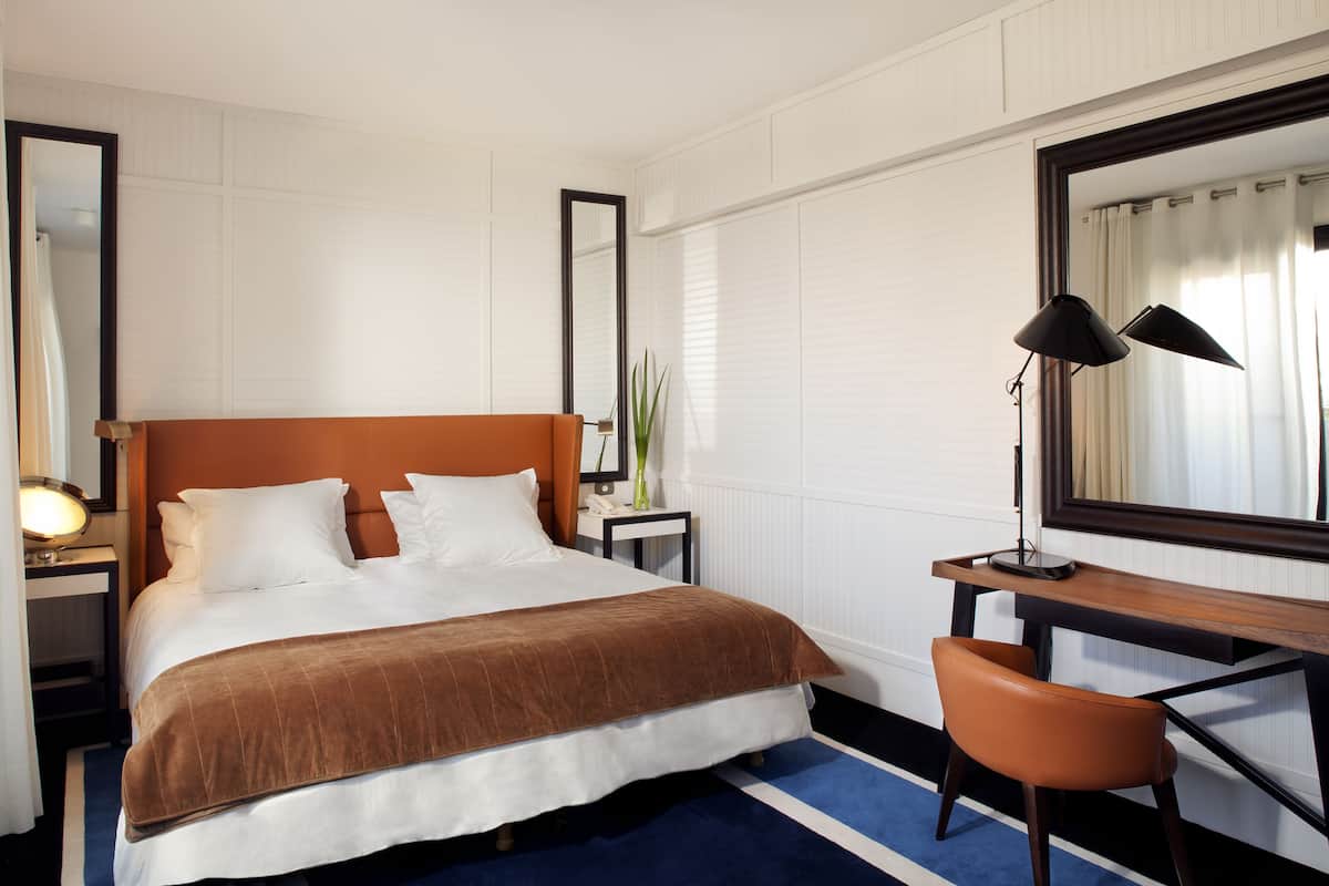 A hotel room with a bed, a desk, a chair, a mirror, and a lamp.