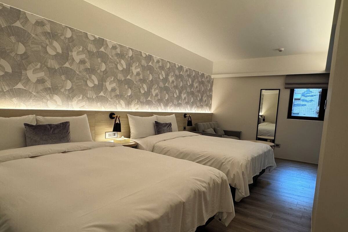 A modern hotel room with two beds, a large mirror, and a view of the city through a window.