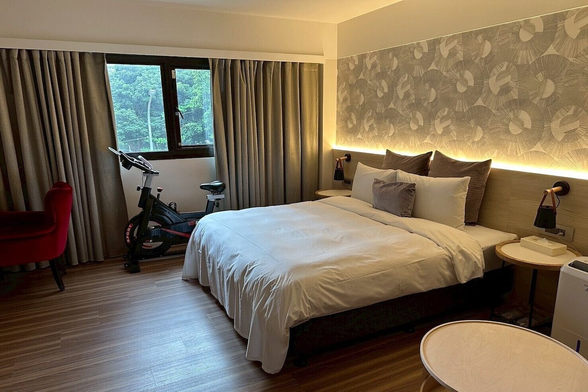 A hotel room with a bed, a bicycle, a red chair, a bedside table, and a window with curtains.